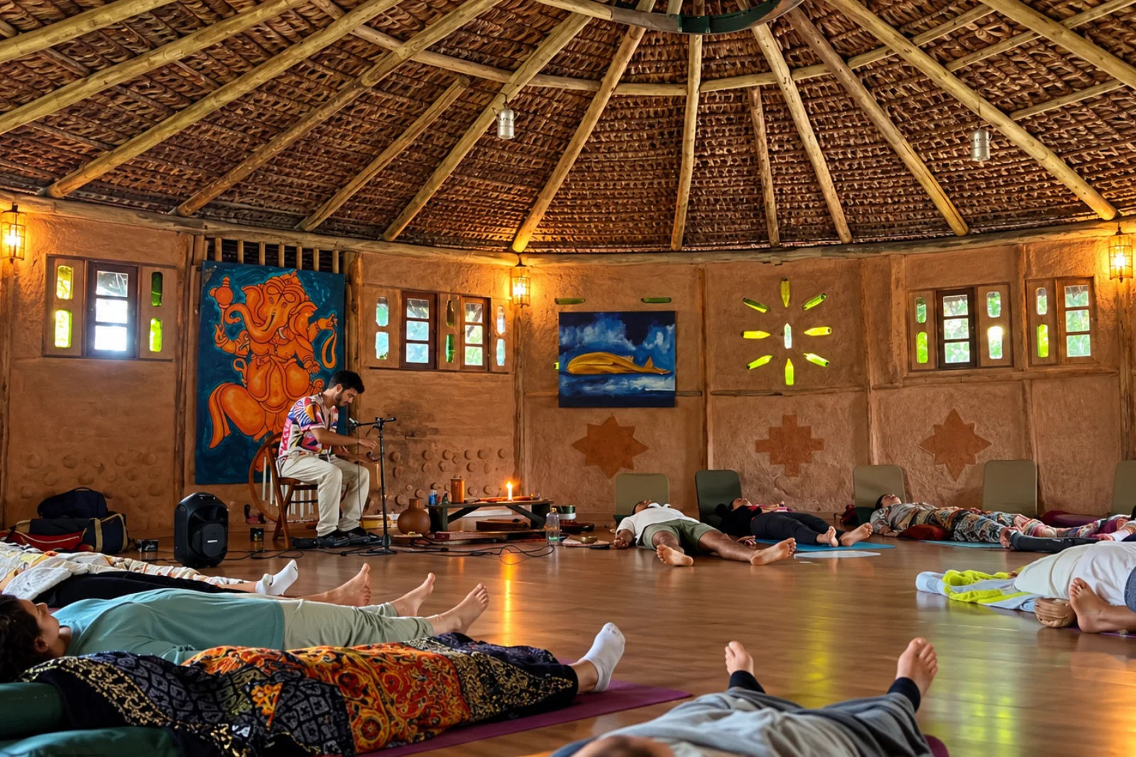 Vivência de Sound Healing durante um retiro de yoga no Espaço Caapora, no Campeche, em Florianópolis, com participantes deitados em colchonetes em um ambiente circular de madeira, teto de palha e paredes de terra.